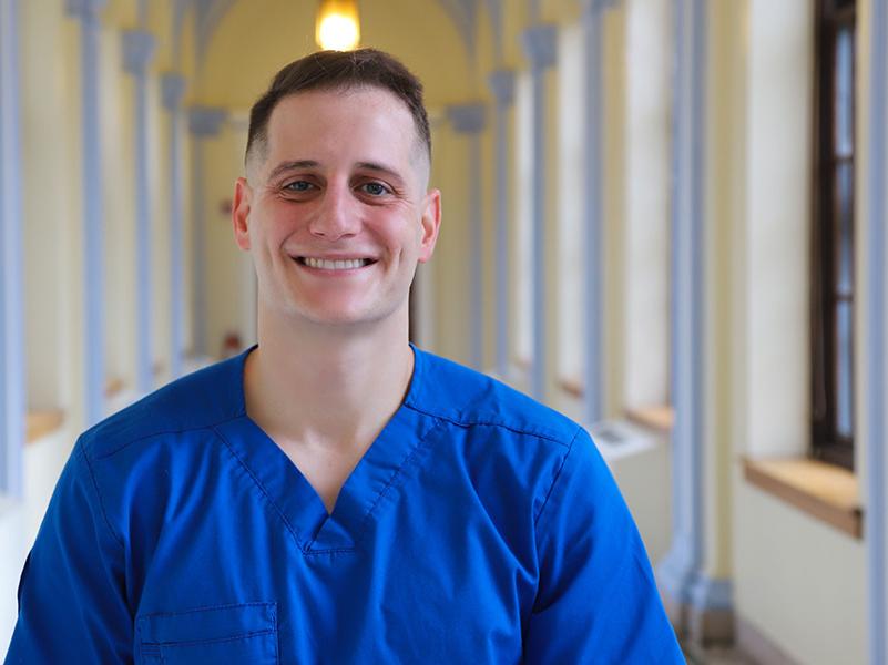 Man in hallway, wearing blue scrubs.